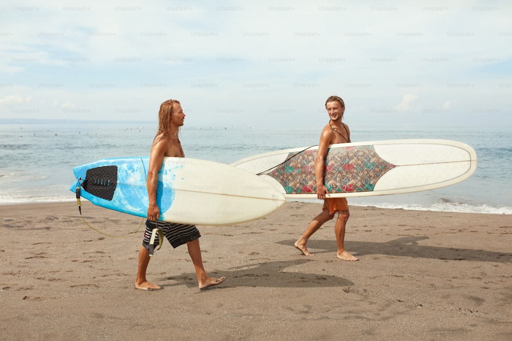 Surf. Giovani surfisti con tavole da surf. Uomini belli sorridenti che camminano sulla spiaggia dell'oceano. Stile di vita attivo, sport acquatici su uno splendido sfondo marino.