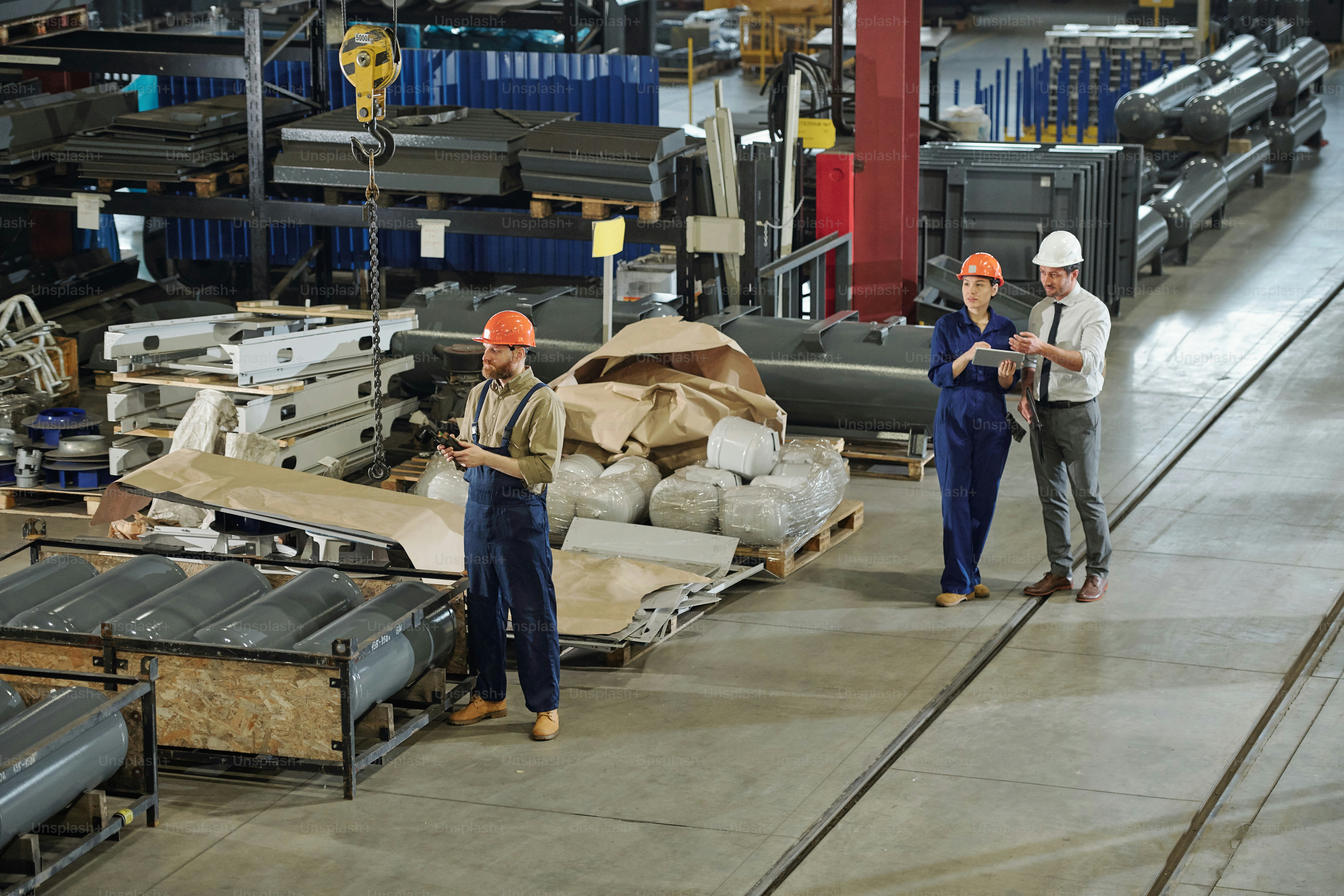People working at factory warehouse: bearded man using industrial crane with remote control while his colleagues discussing products
