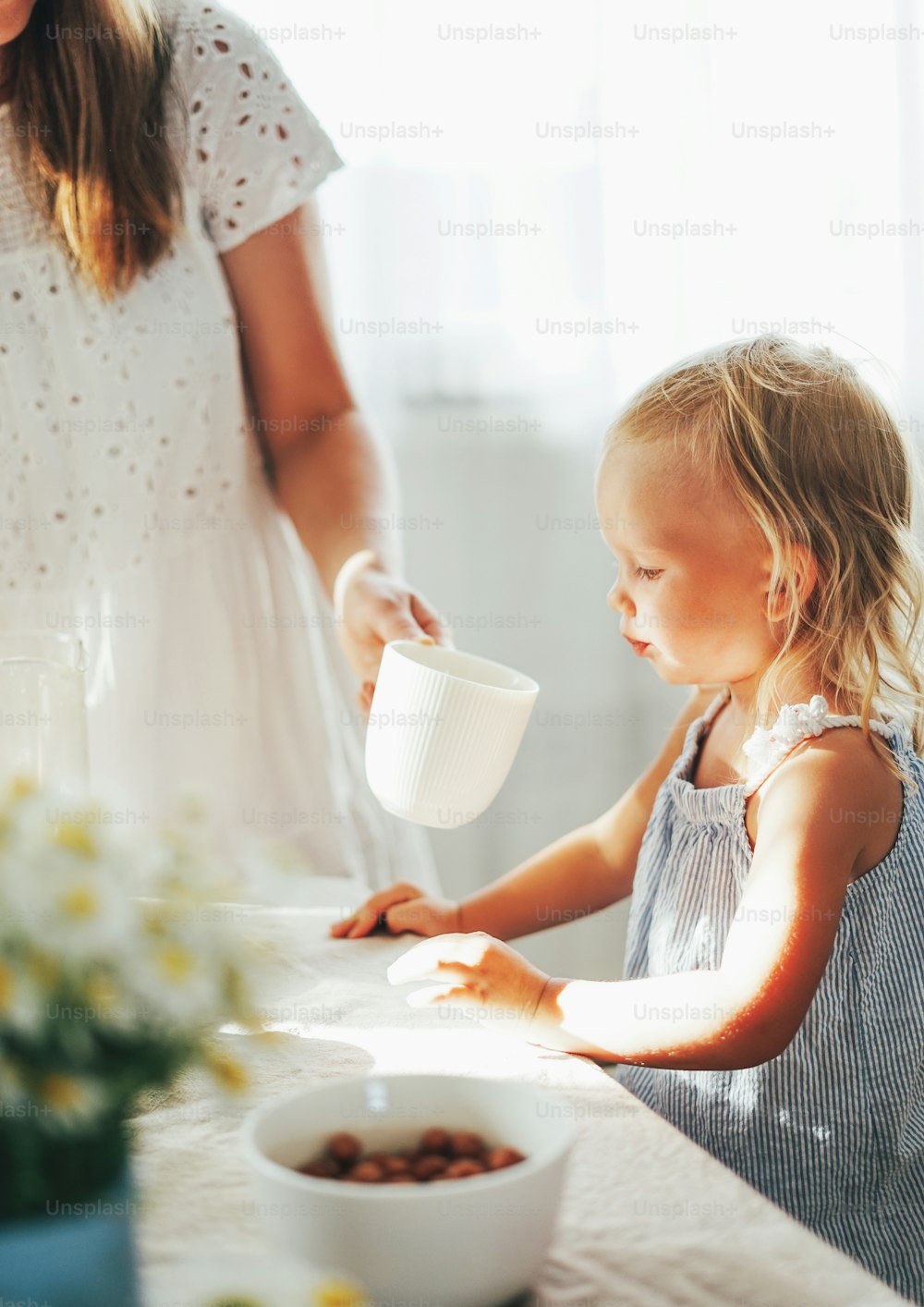 Cute blonde toddler girl drink from cup from mother's hand at table on sunny day. Cottagecore Aesthetics