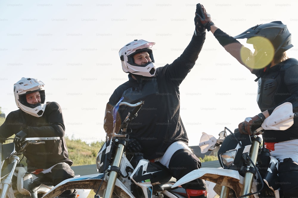 Positive excited men sitting on motorcycles and giving high five while supporting each other before race outdoors