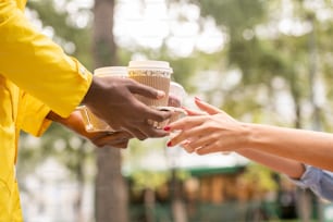 노란색 유니폼을 입은 남성 택배의 손이 두 잔의 커피와 용기에 음식을 담고 있는 식당이나 카페에서 공원에 있는 젊은 여성에게 전달합니다.
