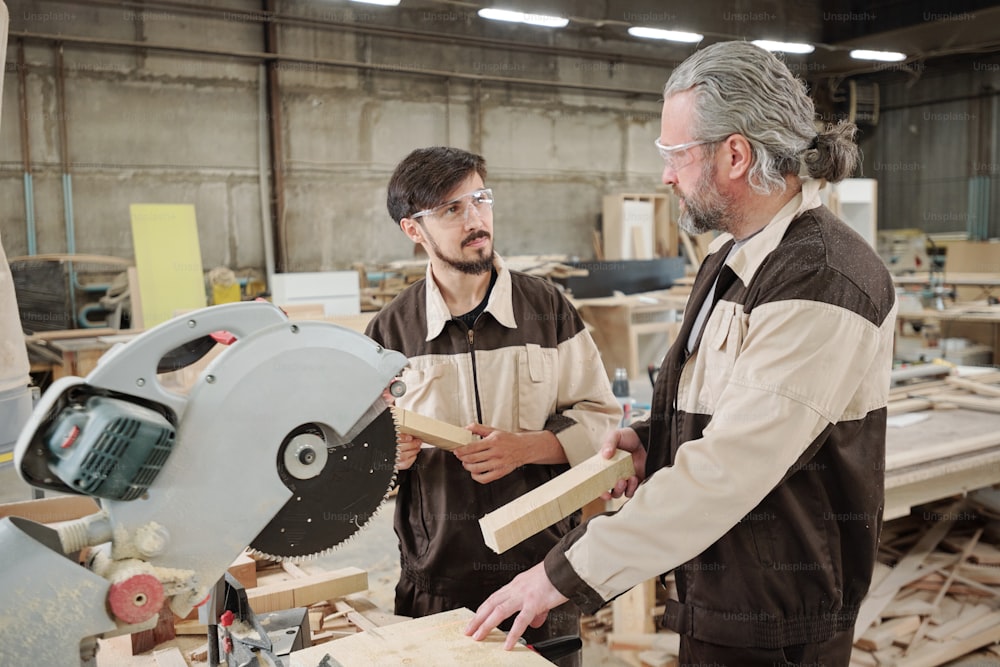 Mature factory worker in workwear looking at young colleague while consulting him about how to use electric circle saw