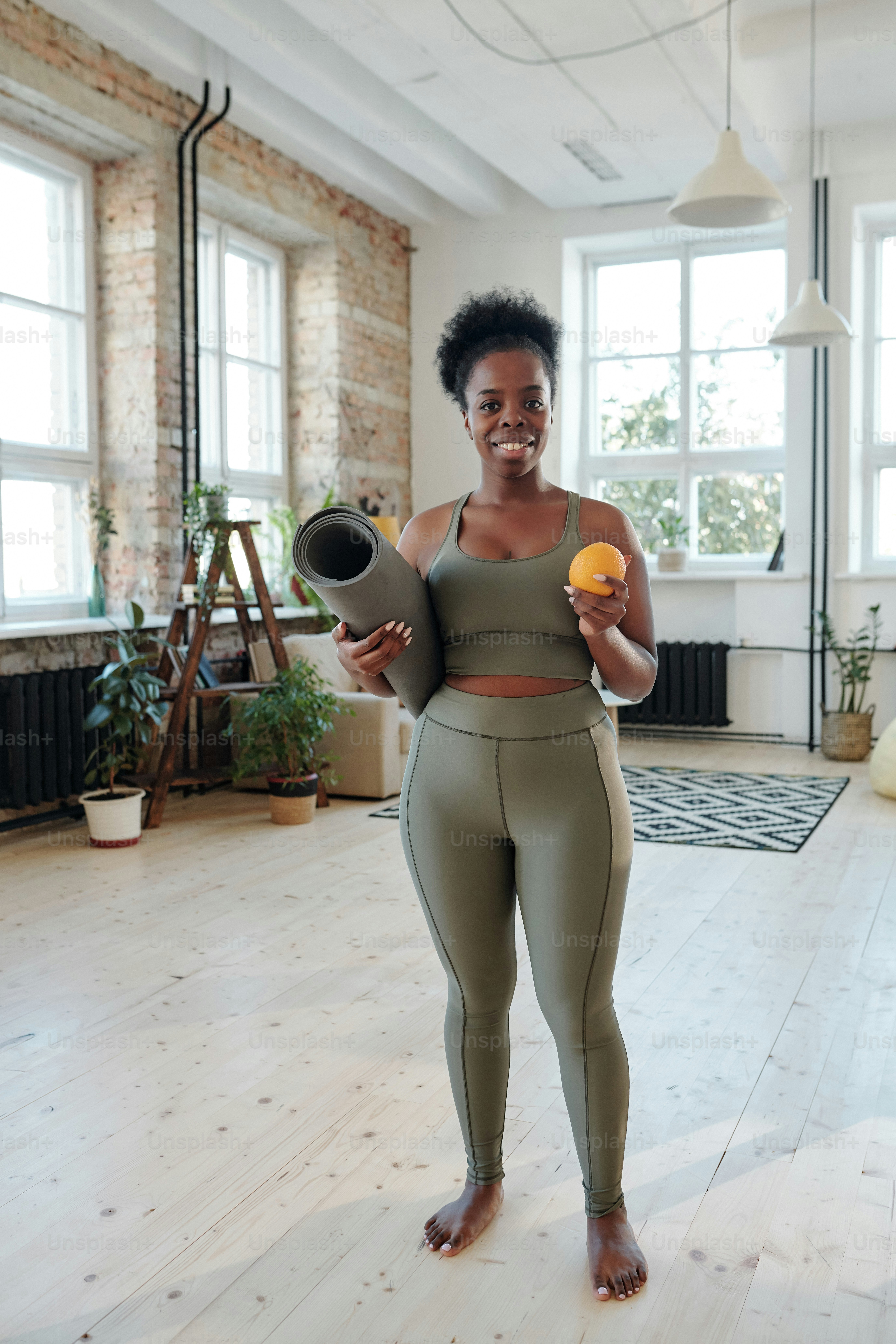 Giovane donna africana sana in forma che ti guarda con un sorriso a denti stretti mentre va ad allenarsi sul pavimento del soggiorno a tuo piacimento
