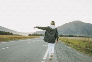 Blonde young woman traveler in white hoodie on road, trip to mountains, hitching ride