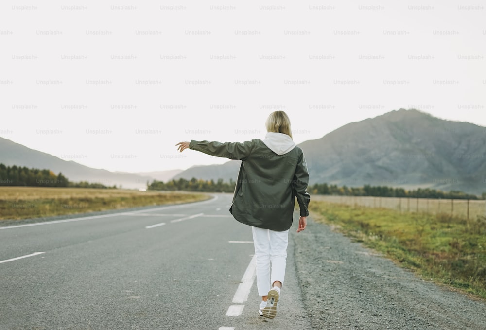 Blonde young woman traveler in white hoodie on road, trip to mountains, hitching ride