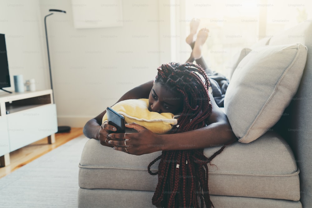 긴 머리띠를 가진 아름다운 젊은 흑인 여성은 소파에서 휴식을 취하고 스마트폰을 통해 친구들과 이야기하고 있습니다. 졸린 아프리카 여성이 집에서 소파에 누워 휴대폰을 사용하여 채팅하고 있다