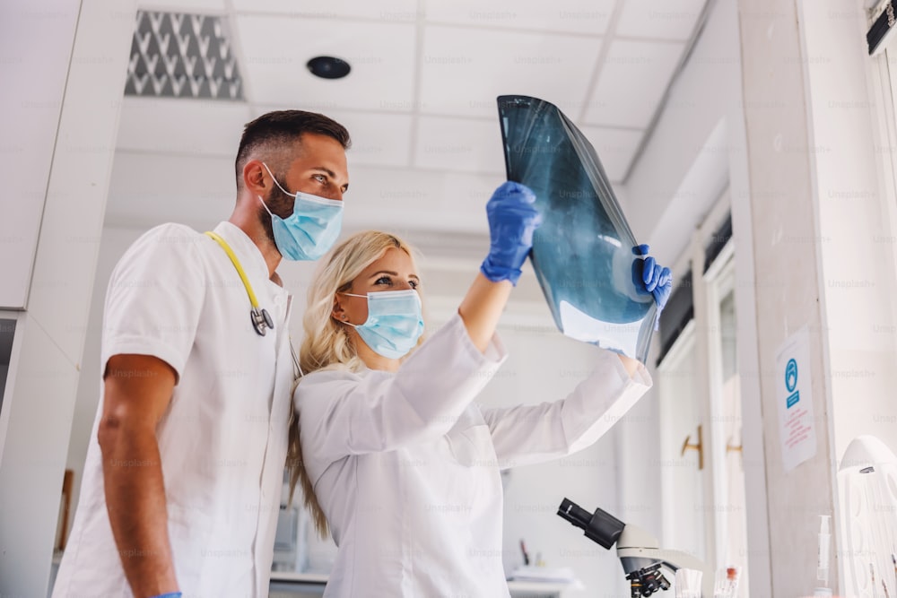 안면 마스크와 고무 장갑을 낀 두 명의 헌신적인 의사가 폐의 엑스레이를 보고 있습니다. 코로나 바이러스 발생 개념.