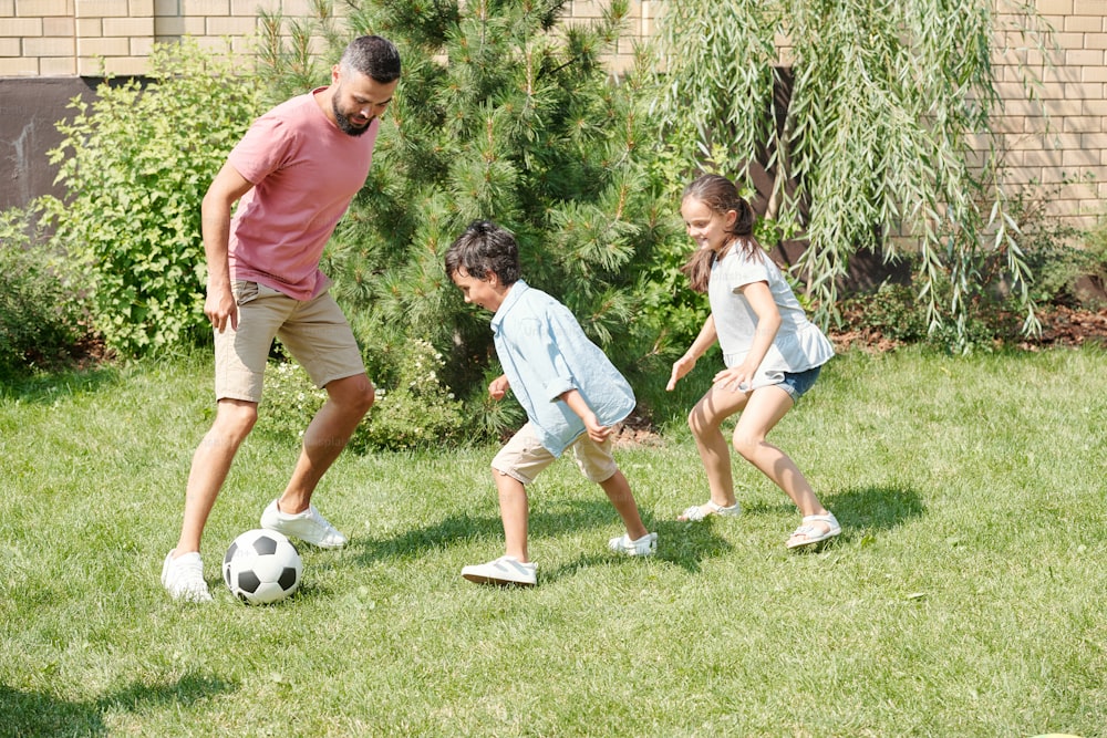 Modern father playing football with his son and daughter on lawn in backyard