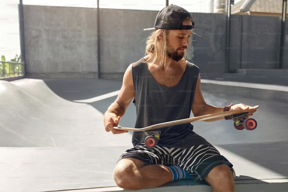 Skater man at skatepark. portrait of handsome guy in casual outfit with  skateboard sitting on concrete ramp. hipster with extreme sport equipment  at park. skateboarding as urban lifestyle. photo – Casual clothing