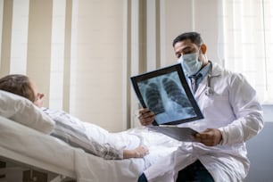 Young radiologist in whitecoat and protective mask looking at lung x-ray image of sick female patient while sitting by her bed in chamber
