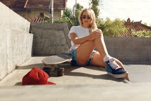 Subkultur. Skater-Mädchen sitzt auf Betonrampe im Skatepark. Teenager in lässigem Outfit mit modernen Sportgeräten posiert im Freien. Urbaner Lebensstil für aktive Teenager in der Stadt.