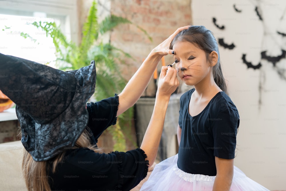 Animador infantil em withches fantasia fazendo maquiagem gatinho para menina asiática na festa de Halloween