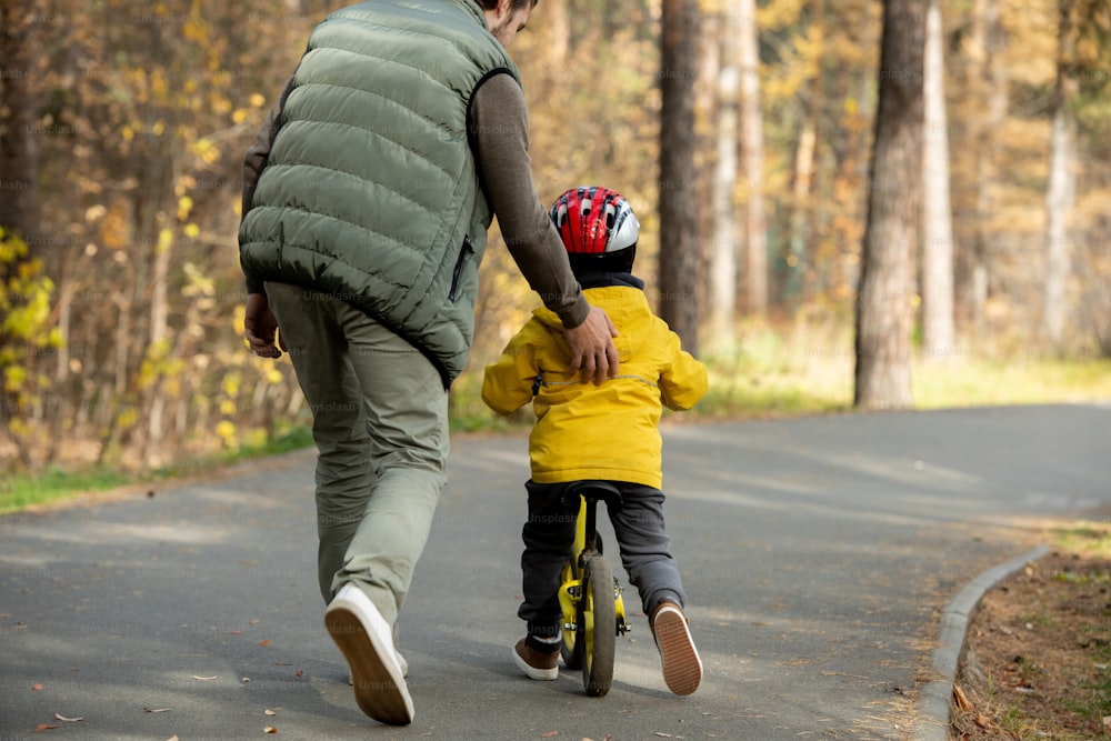 공원에서 균형 자전거를 타는 방법을 가르치면서 안전 헬멧과 캐주얼웨어를 입은 어린 아들을 돕는 젊은 아버지의 뒷모습