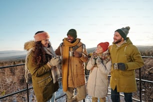 Happy intercultural friends in winterwear having hot tea during chill while standing against blue sky over mountains and forest covered with snow