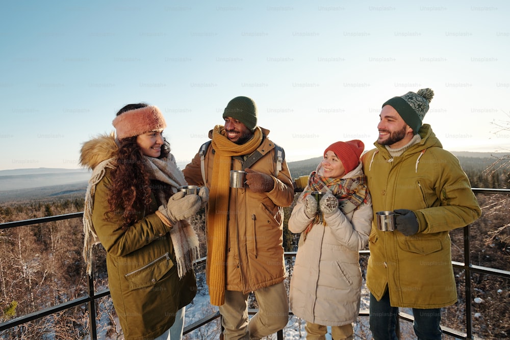 Happy intercultural friends in winterwear having hot tea during chill while standing against blue sky over mountains and forest covered with snow