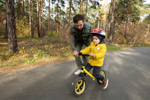 젊은 아버지는 안전 헬멧과 캐주얼웨어를 입은 어린 아들이 공원의 아스팔트 도로를 따라 균형 잡힌 자전거를 타는 것을 도우면서 약간 구부렸다