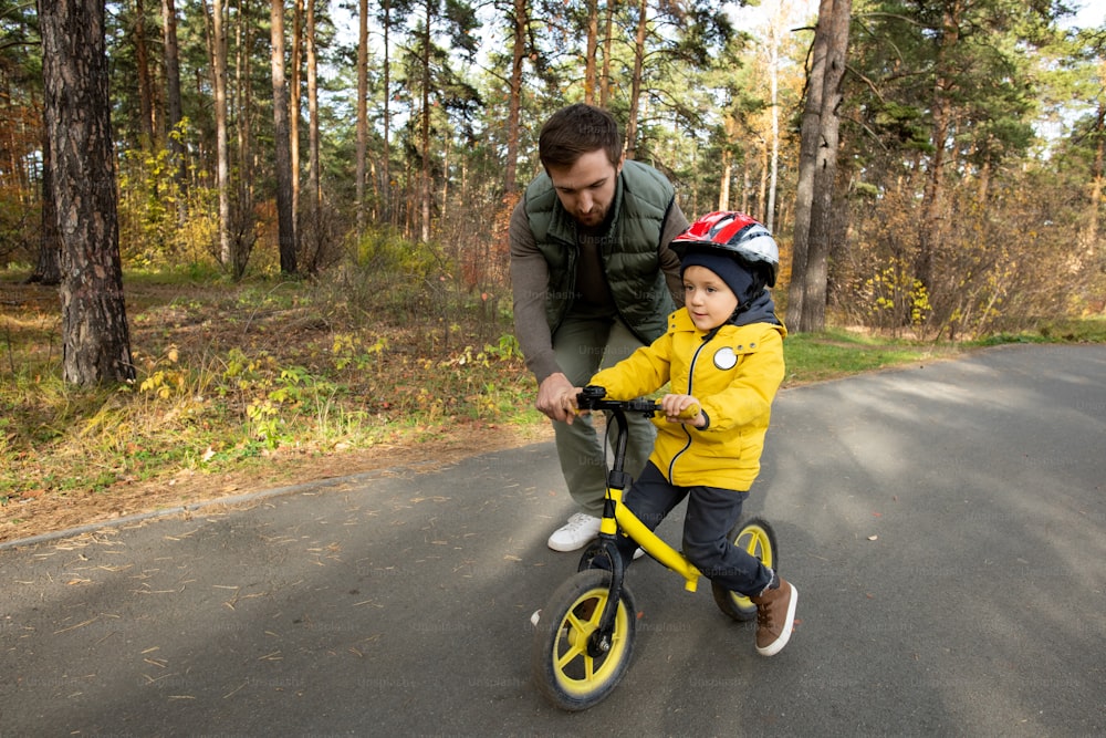 Young father slightly bending while helping his little son in safety helmet and casualwear to ride balance bicycle along asphalt road in park