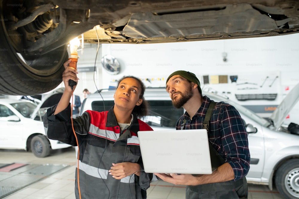 Young female mechanic with lamp and her male colleague with laptop looking at bottom of broken car to find out the reason of problem