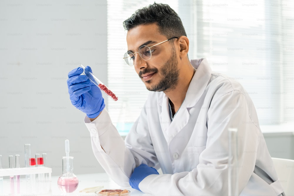 Ricercatore contemporaneo guantato in camice bianco che guarda la fiaschetta con diversi piccoli campioni di carne vegetale cruda durante un esperimento scientifico in laboratorio