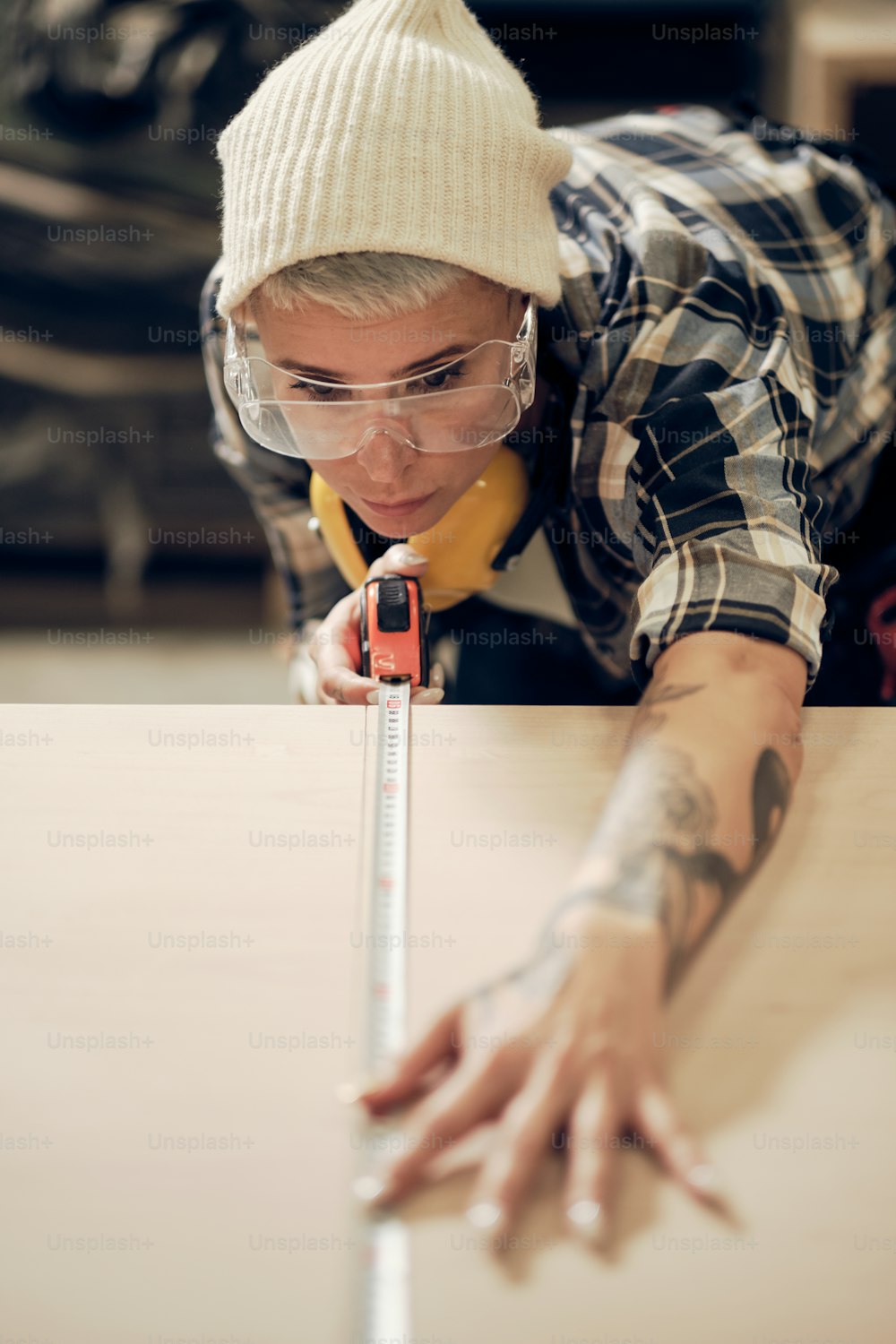 Vertical portrait of tattooed blond woman working at wood workshop, measuring with tape, concentrated