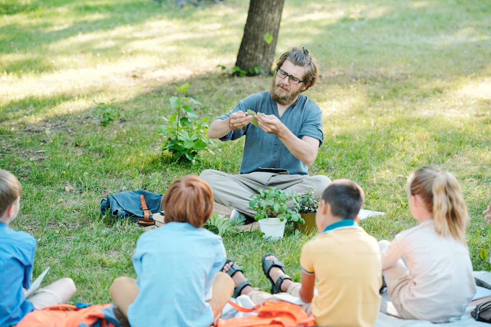 Young botany teacher in glasses holding plant leaf and explaining photosynthesis process to pupils at outdoor class