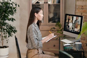 Side view of young female office manager sitting by table in front of computer monitor and making notes while talking to people on screen