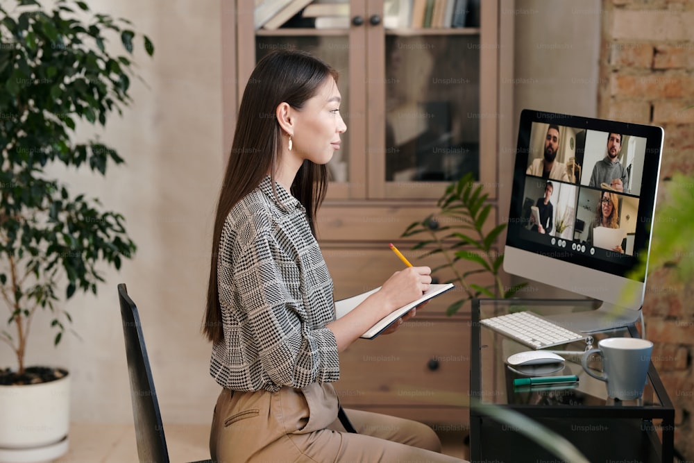 Side view of young female office manager sitting by table in front of computer monitor and making notes while talking to people on screen