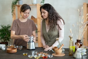 Young woman using electric kitchen appliance for grinding ingredients for handmade soap and showng her friend which button to press