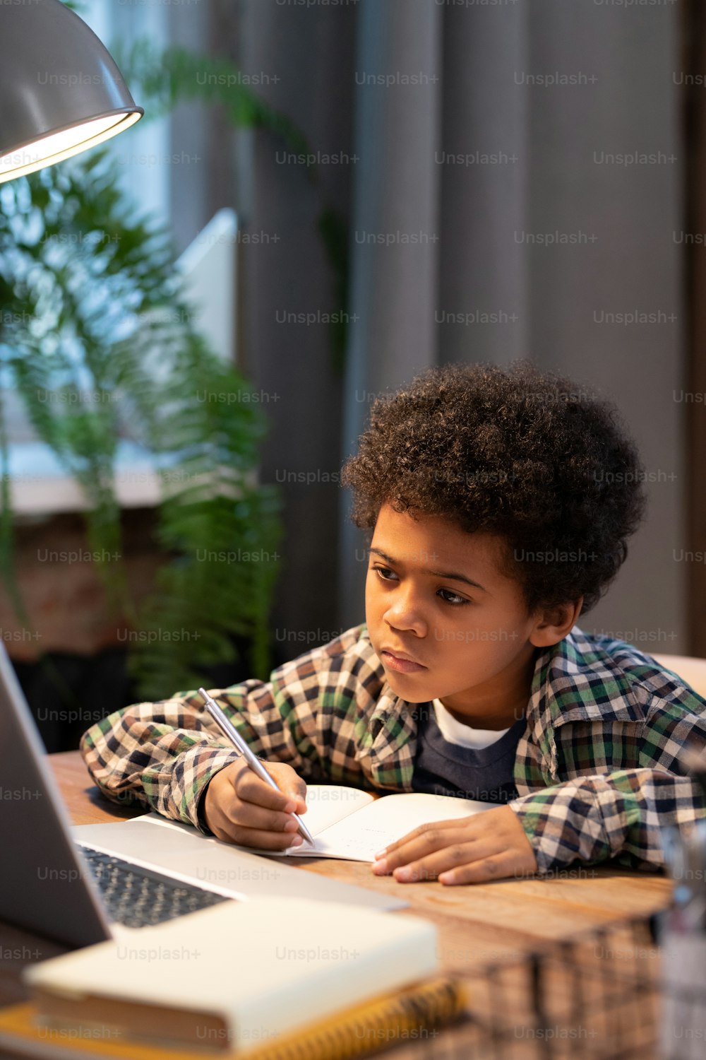Serious African schoolboy making notes in copybook while sitting by table in front of laptop and looking at display during online lesson