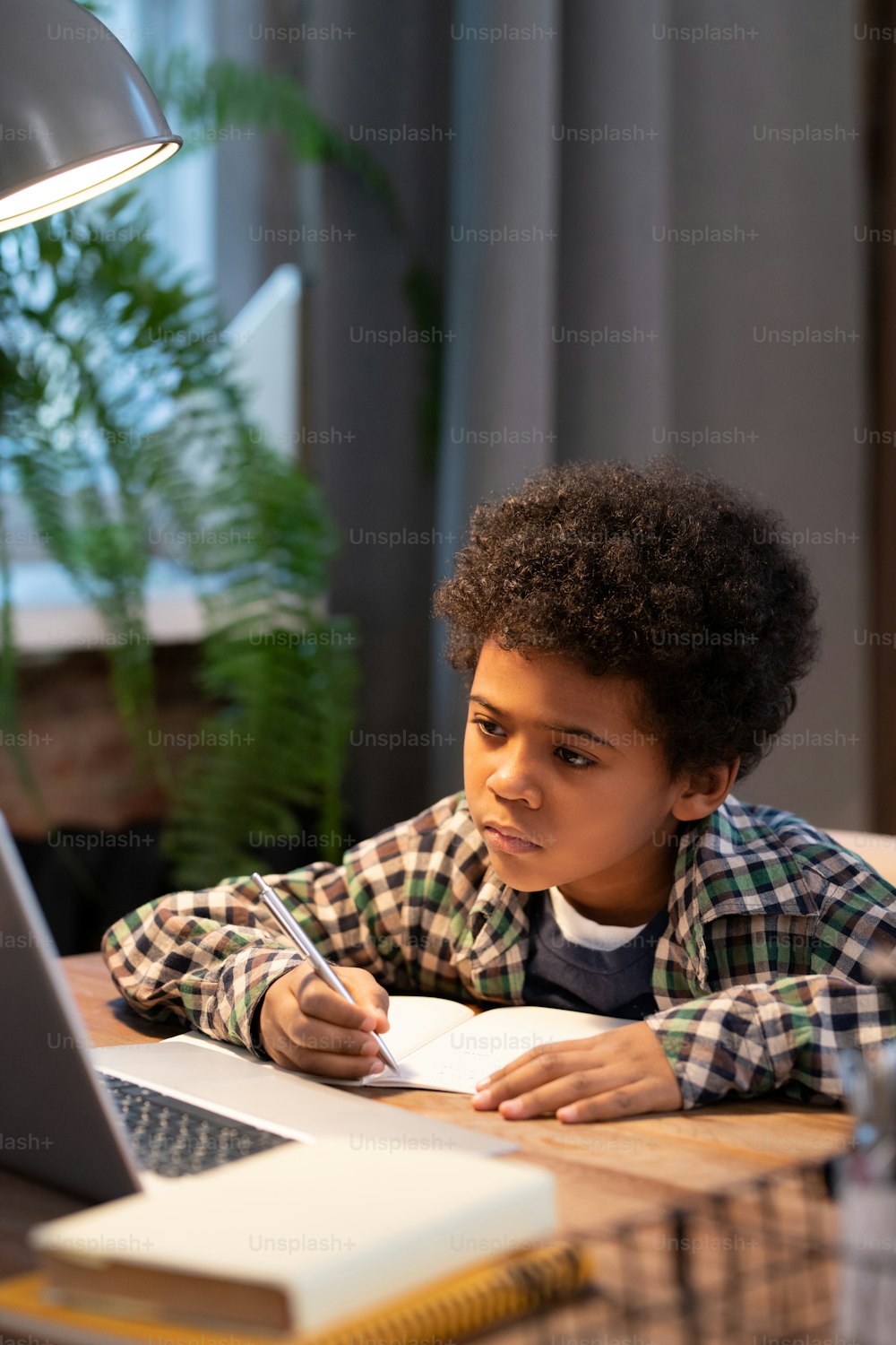 Ernsthafter afrikanischer Schüler, der Notizen im Copybook macht, während er am Tisch vor dem Laptop sitzt und während des Online-Unterrichts auf das Display schaut