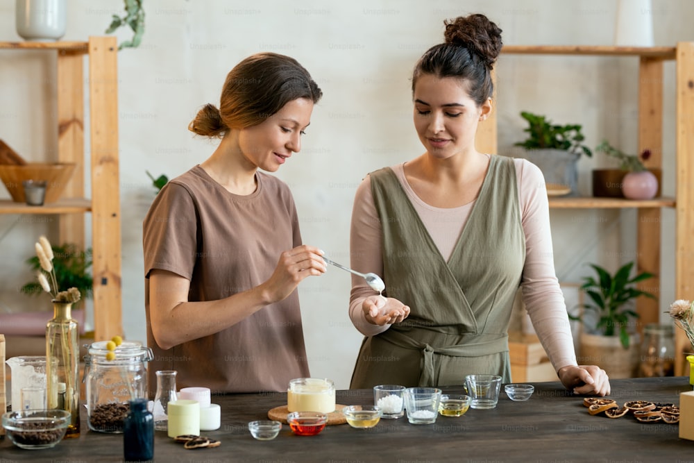 Young female putting blob of fresh handmade liquid soap mass on palm of her friend helping her with making natural handmade cosmetic products