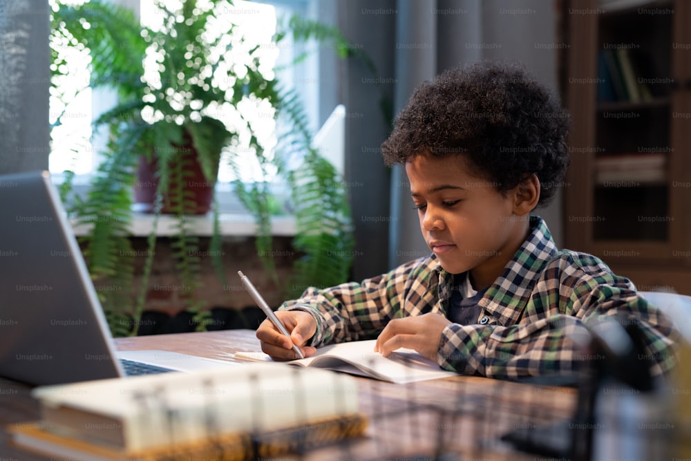캐주얼 복장을 입은 귀여운 아프리카 남학생이 노트북 앞 테이블에 앉아 가정 과제를 수행하는 동안 카피북에 메모를 하고 있다