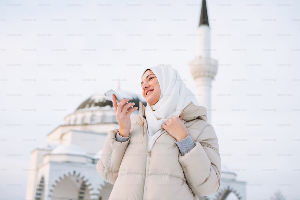 Beautiful smiling young Muslim woman in headscarf in light clothing using mobile against the background of mosque in winter season