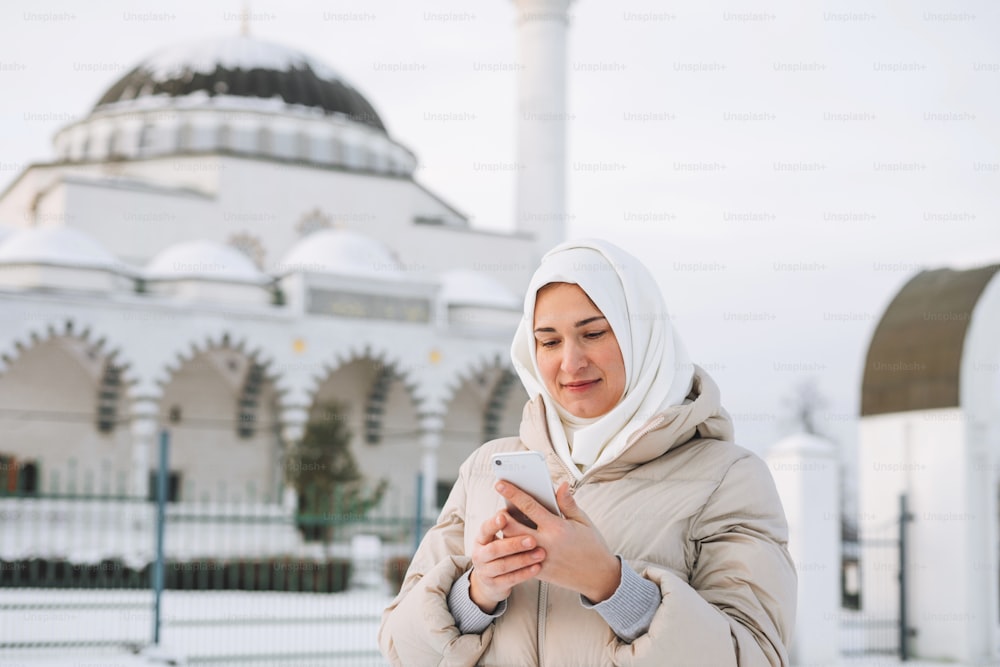 Beautiful smiling young Muslim woman in headscarf in light clothing using mobile against the background of mosque in winter season