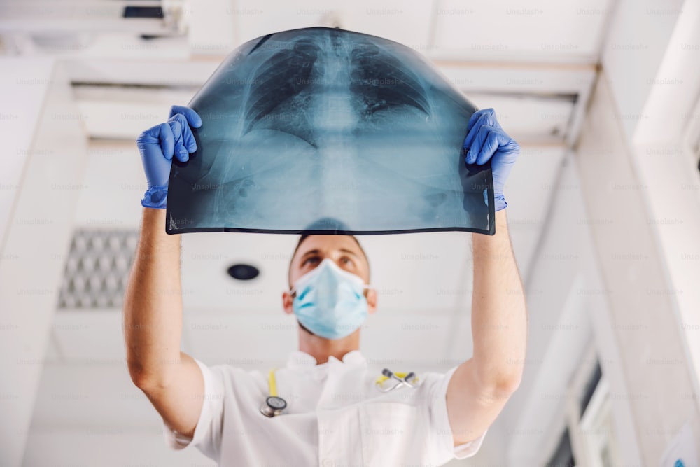 Arzt mit Gesichtsmaske und Gummihandschuhen, der im Krankenhaus steht und sich die Röntgenaufnahme der Lunge während des Coronavirus ansieht.