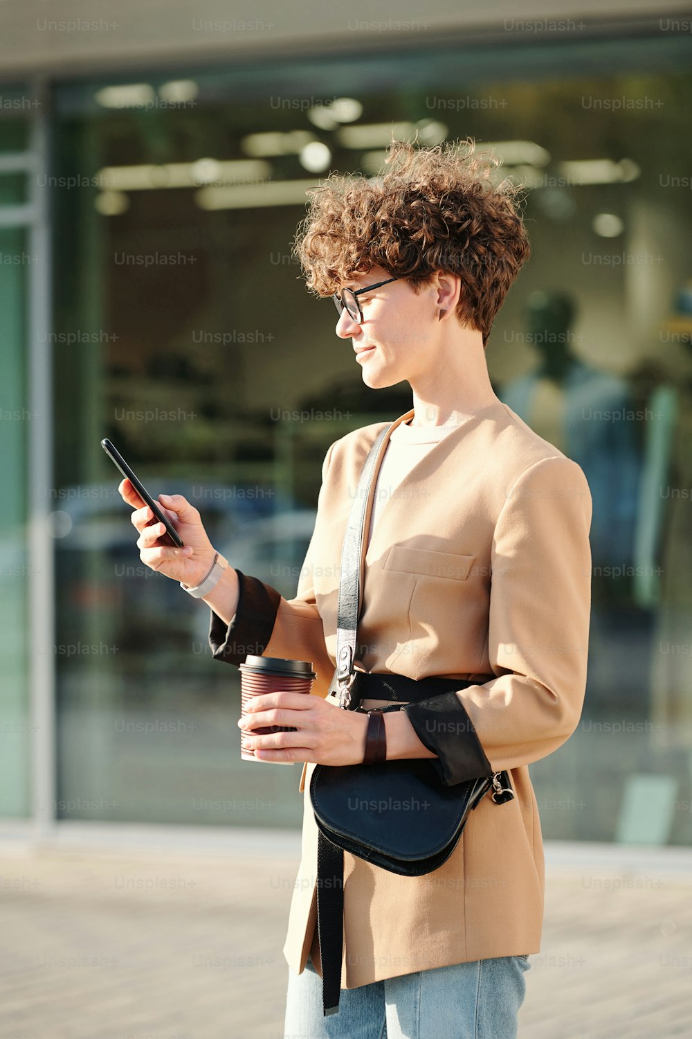 Junge brünette Geschäftsfrau mit Getränk und Handtasche SMS im Smartphone, während sie gegen das große Schaufenster des Einkaufszentrums steht