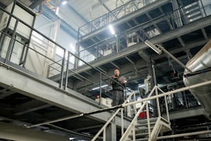 Bald mature foreman with digital tablet working inside contemporary large polymer processing factory while standing in front of staircase