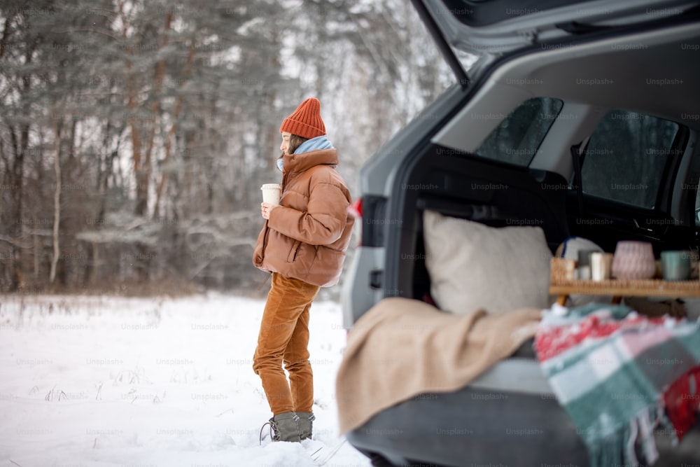 Woman with hot drink staying near car trunk in a pine snowy forest, traveling by car during winter holidays. High quality photo