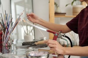 Hands of young female artisan sitting by workplace and burning glass workpiece with fire while holding it over burner in workshop