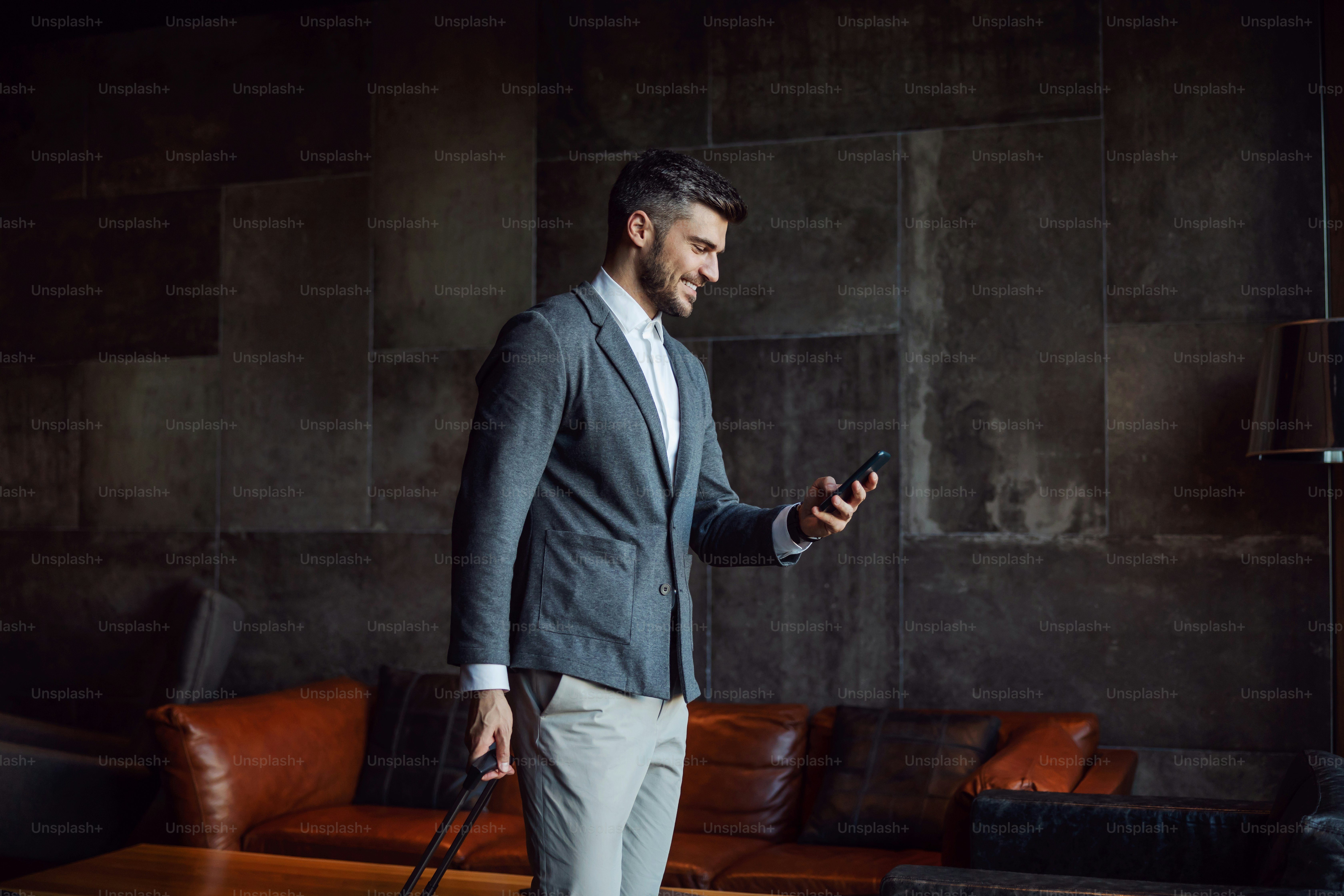 Happy businessman carrying his luggage while walking at the hall of a hotel and using phone. Business trip, travel, technologies