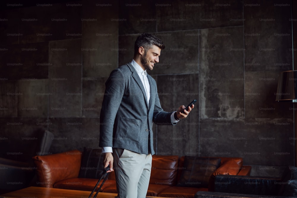 Happy businessman carrying his luggage while walking at the hall of a hotel and using phone. Business trip, travel, technologies