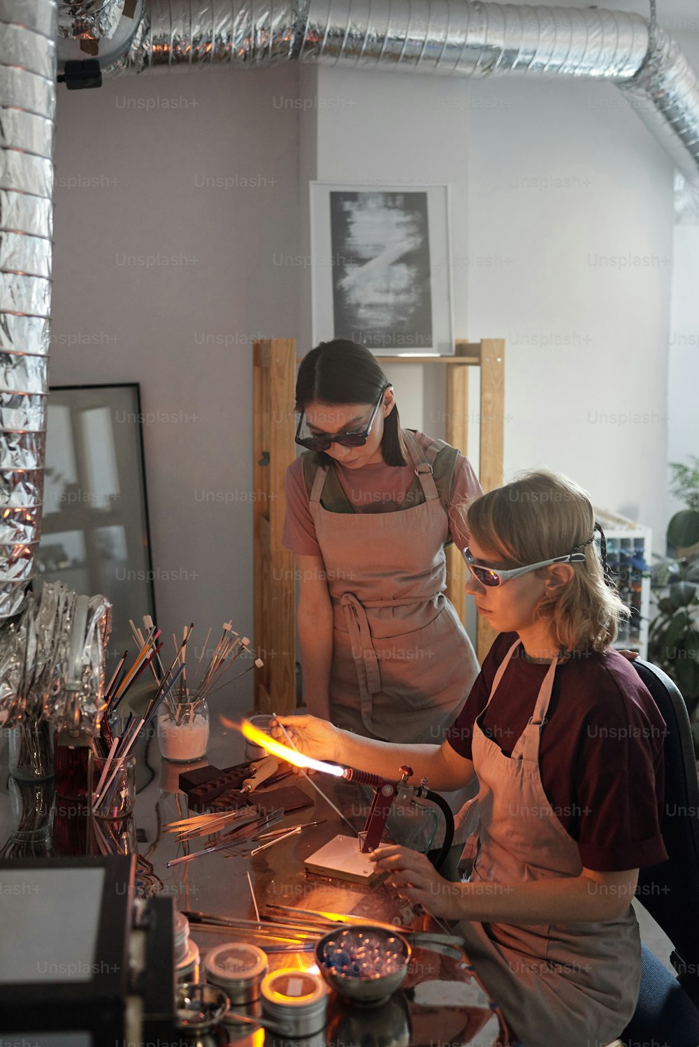 Jovem artesã contemporânea em roupas de trabalho e óculos mostrando a sua estagiária como queimar peças de vidro longas em queimador durante masterclass