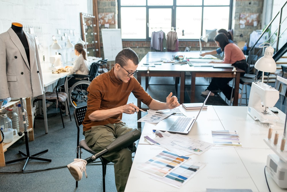 Joven diseñador serio con pierna protésica sentado junto al escritorio frente a la computadora portátil mientras hace bocetos para la nueva colección de temporada