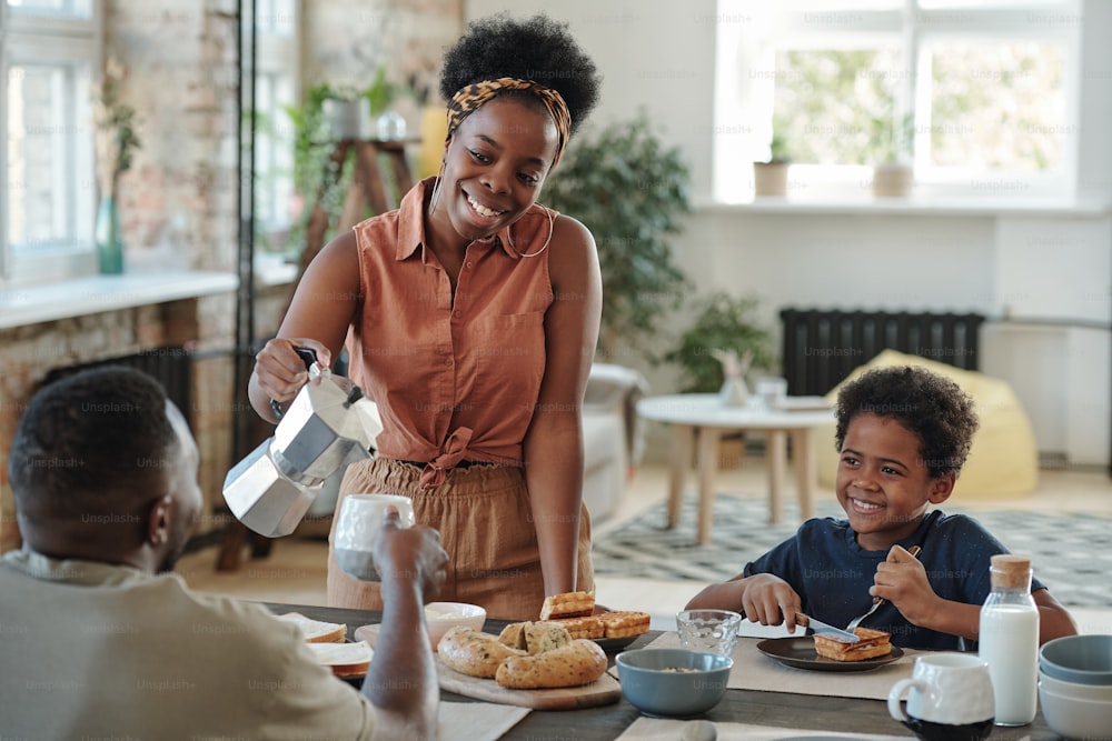 Young happy African woman pouring coffee or tea into mug of her husband while their cute little son having dessert during breakfast