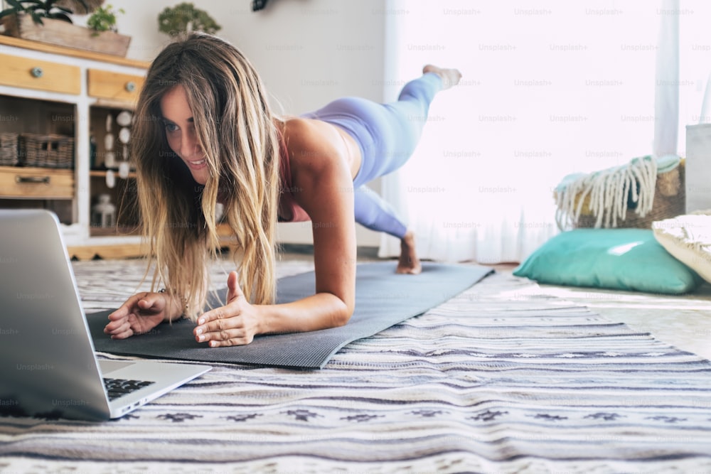 Eine sportliche Frau in Sportbekleidung, die Plank-Pilates-Position auf dem Boden mit einem Laptop-Computer zu Hause im Wohnzimmer macht. Sport- und Erholungskonzept und gesunder Lebensstil Indoor