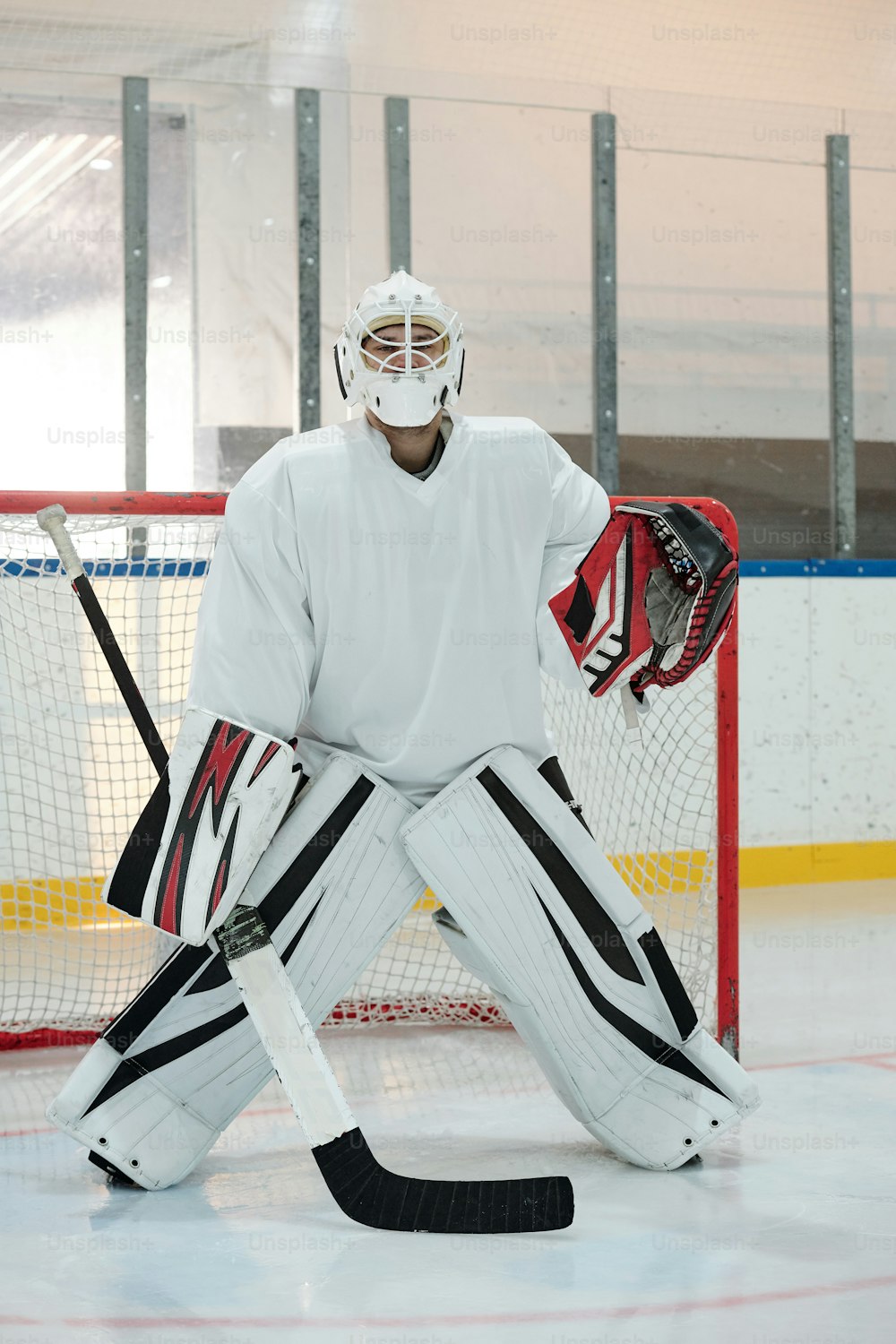 Zeitgenössischer Hockeyspieler in Sportuniform, Schutzhelm und Handschuhen, der den Stock hält, während er auf der Eisbahn gegen das Netz steht und auf den Puck wartet