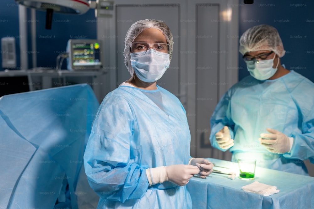 防護マスク、手袋、眼鏡を身に着けたプロの女性外科医が、手術の準備をしている男性の同僚に対してあなたを見つめています