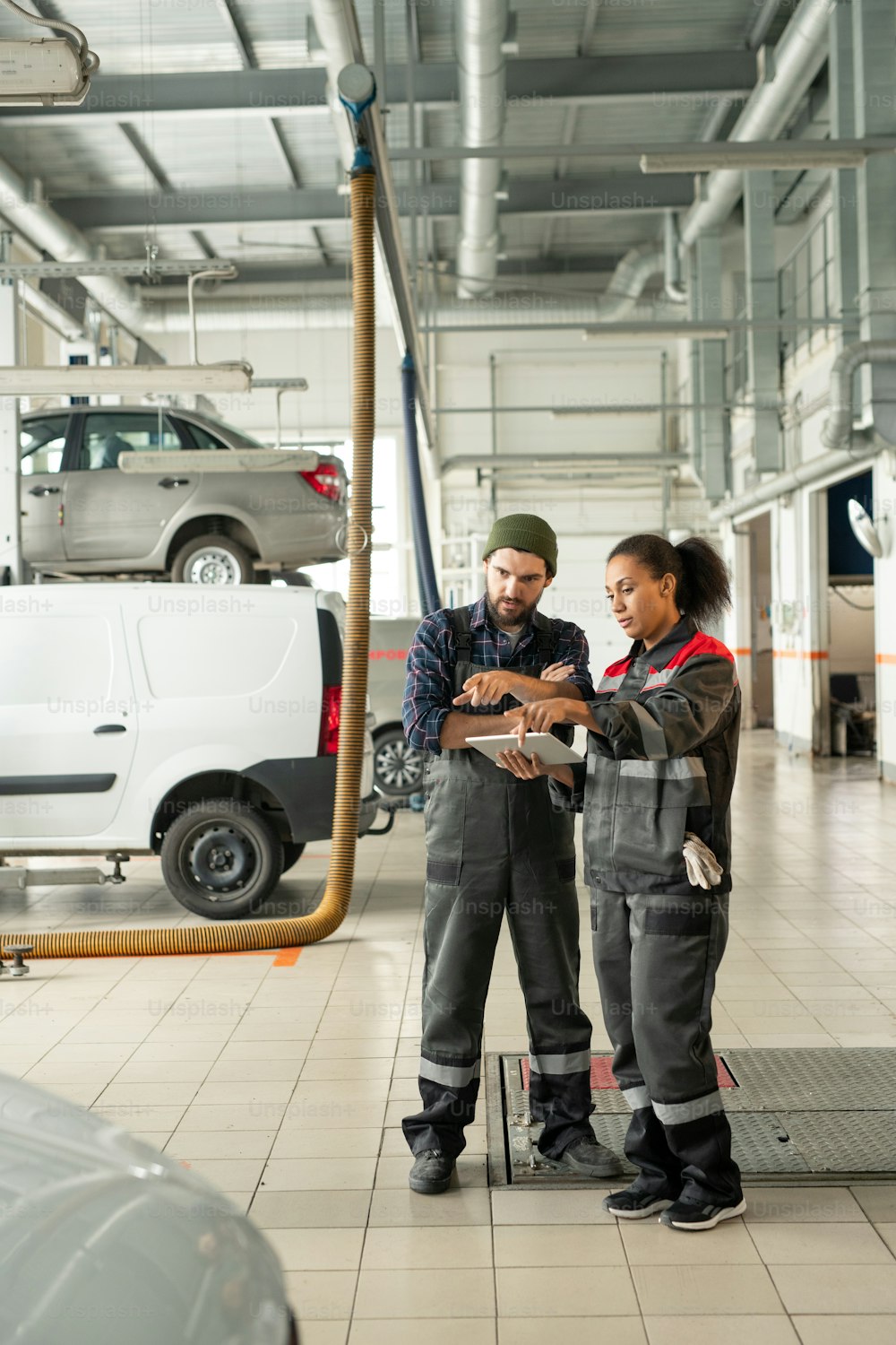 Young confident technicians in workwear pointing at one of cars in workshop while discussing its technical characteristics before repairing it