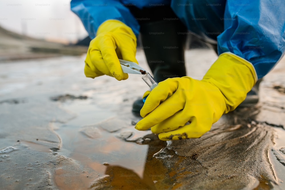 Contemporary ecologist in yellow rubber gloves and protective blue coveralls sitting on squats while taking sample of toxic soil