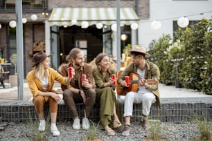 Group of young friends spending summertime together, having fun while sitting in a row with drinks on a porch of the country house